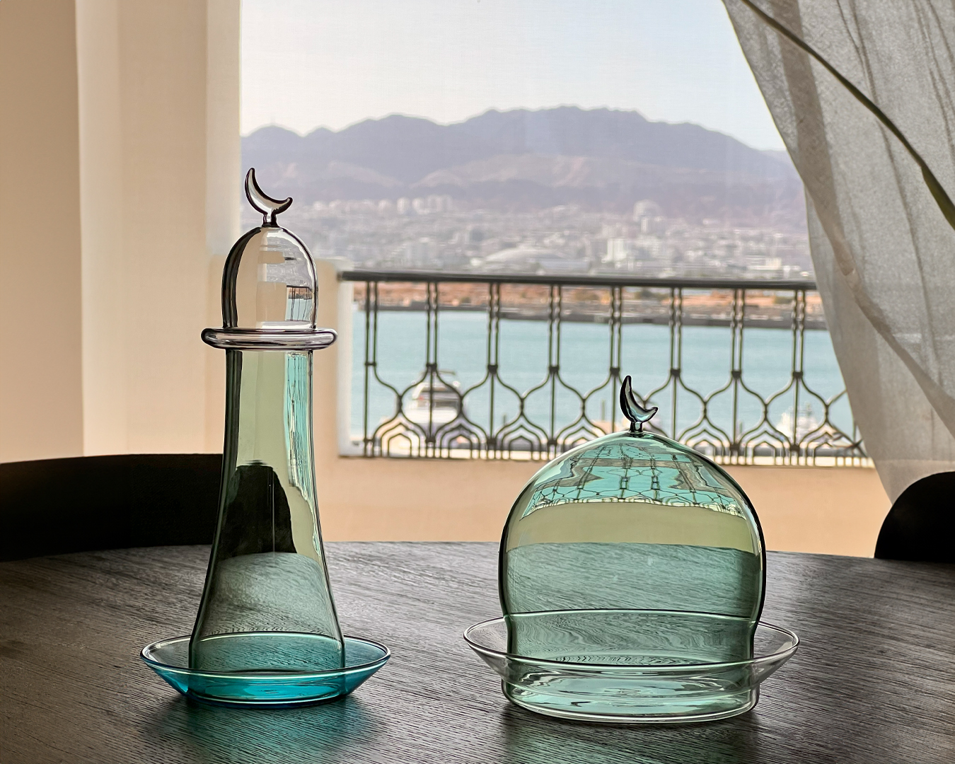 Noor Glass Noor Minaret in Green & Blue, showcased outdoors on a textured surface. The sunlight highlights the intricate details and craftsmanship of the glassware set against a backdrop of a sparkling marina and clear skies.
